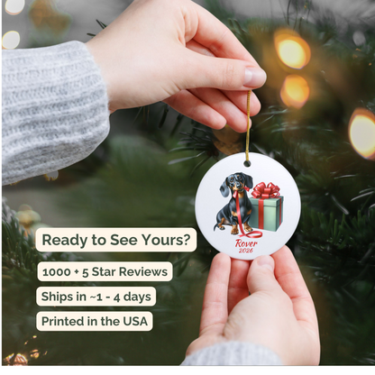 Hand holding a personalized black and tan Dachshund ceramic ornament beside a Christmas tree