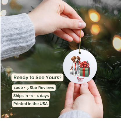 Hand holding a personalized Brittany Spaniel ceramic ornament beside a Christmas tree