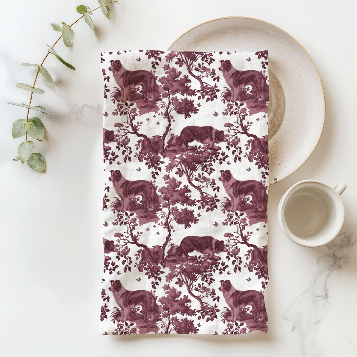 Border Collie Tea Towel with cranberry toile design, folded beside a plate and cup.