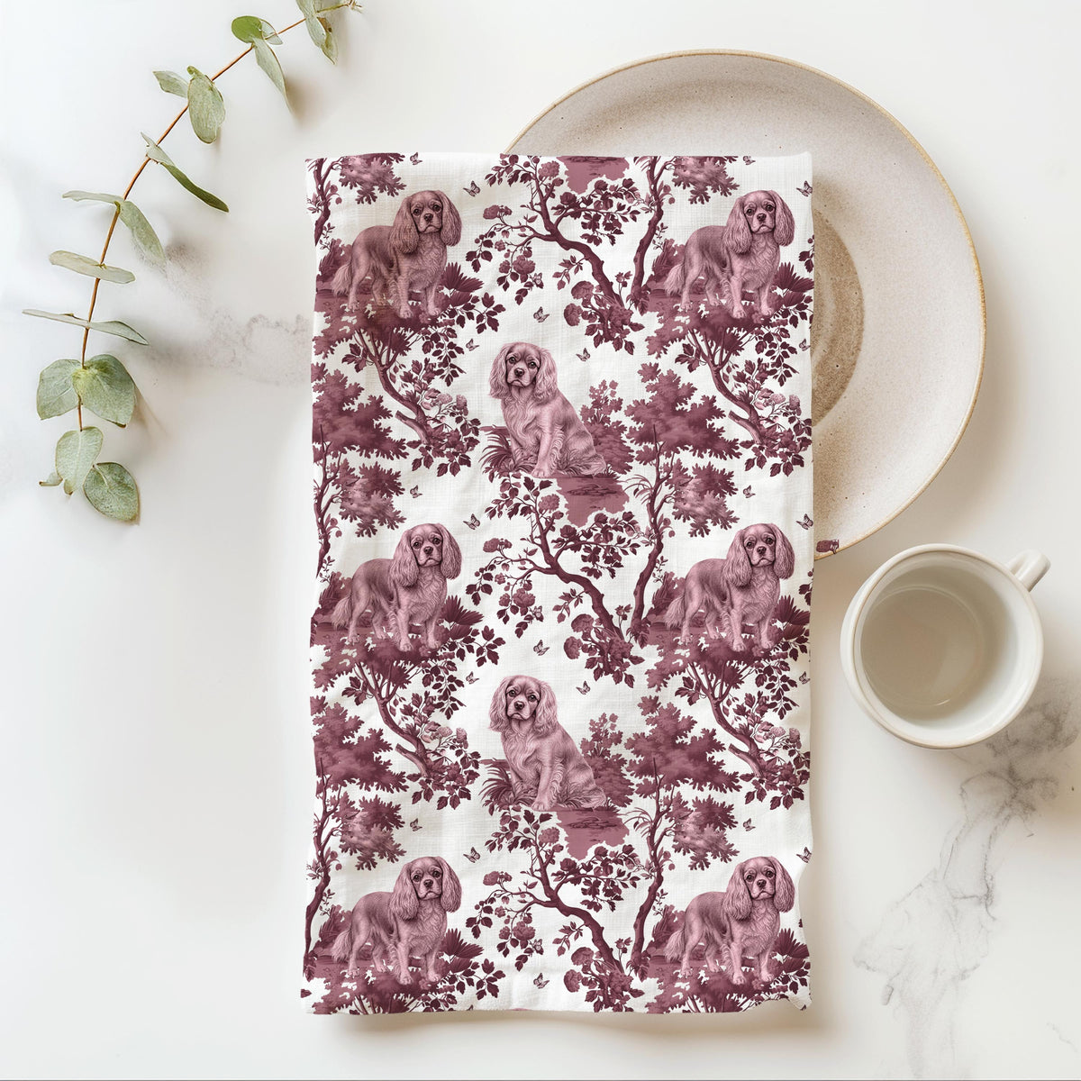 Cavalier King Charles Spaniel Ruby Tea Towel with cranberry toile design, folded beside a plate and cup.