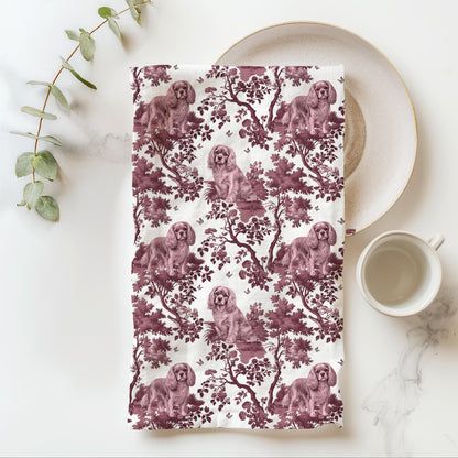 Cavalier King Charles Spaniel Ruby Tea Towel with cranberry toile design, folded beside a plate and cup.