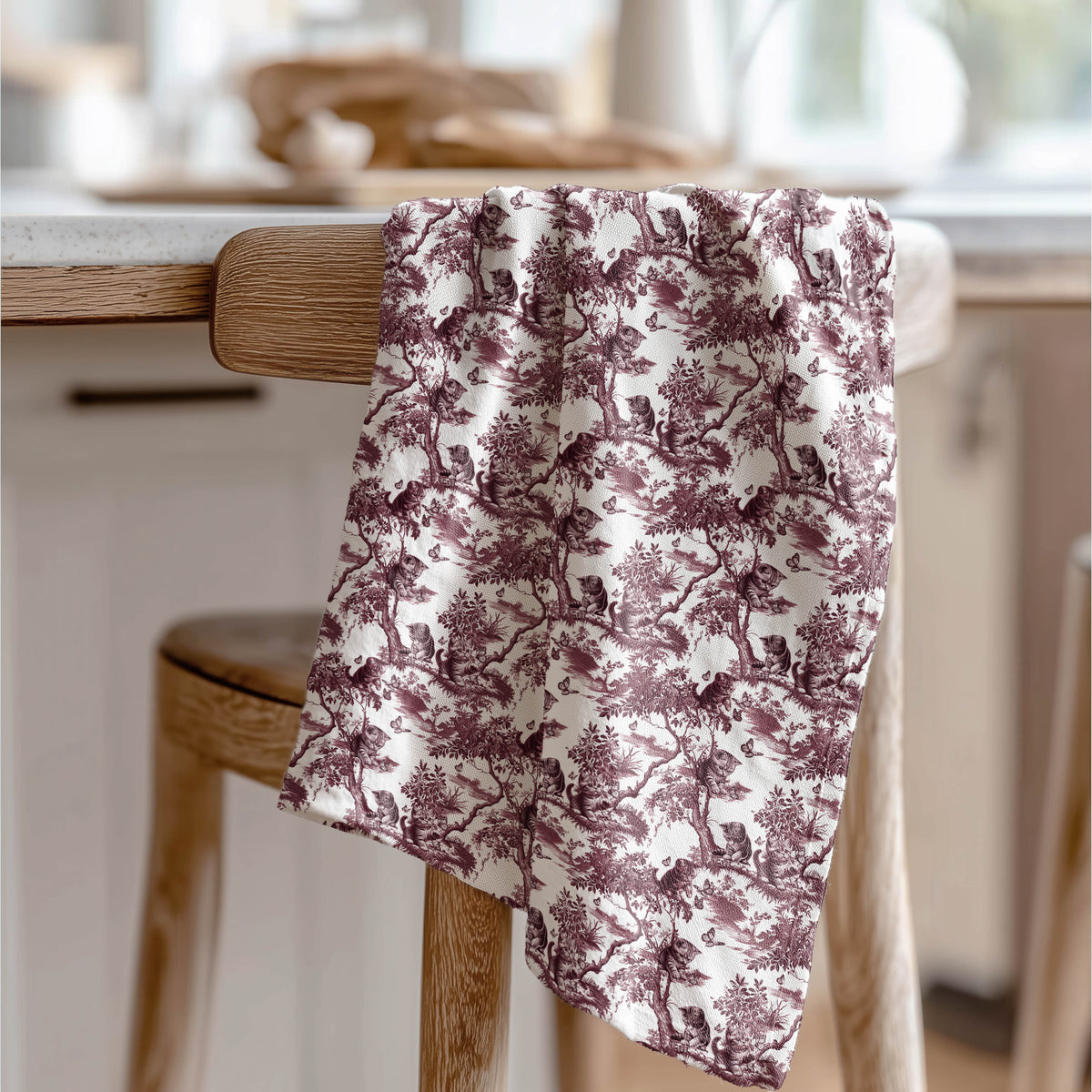 Cat & Butterfly Tea Towel with cranberry toile design, draped over a wooden chair in a kitchen.