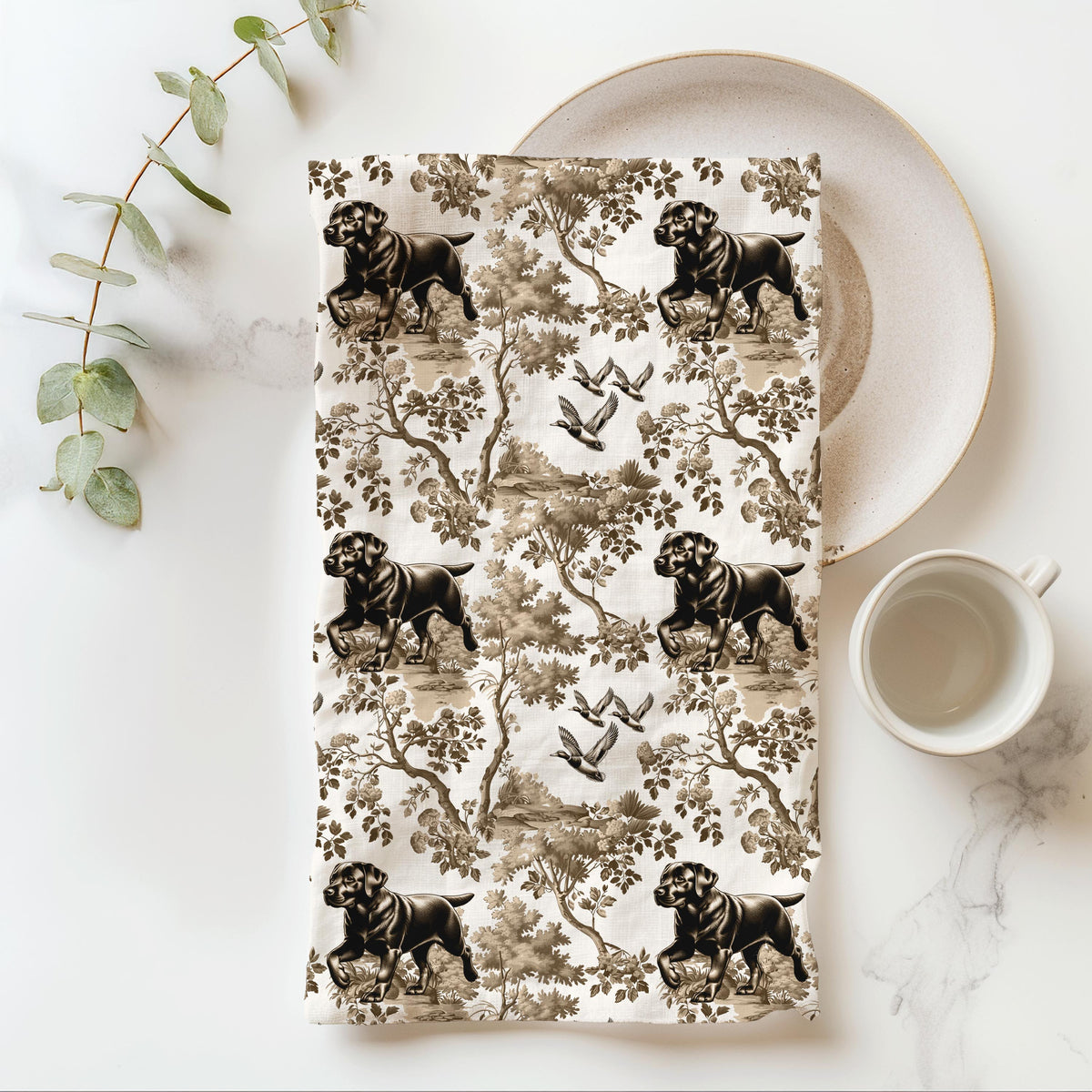 Black Labrador Tea Towel with brown and tan toile design, folded beside a plate and cup.