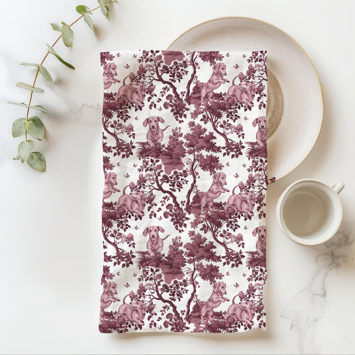 Dachshund Tea Towel with cranberry toile design, folded beside a plate and cup.