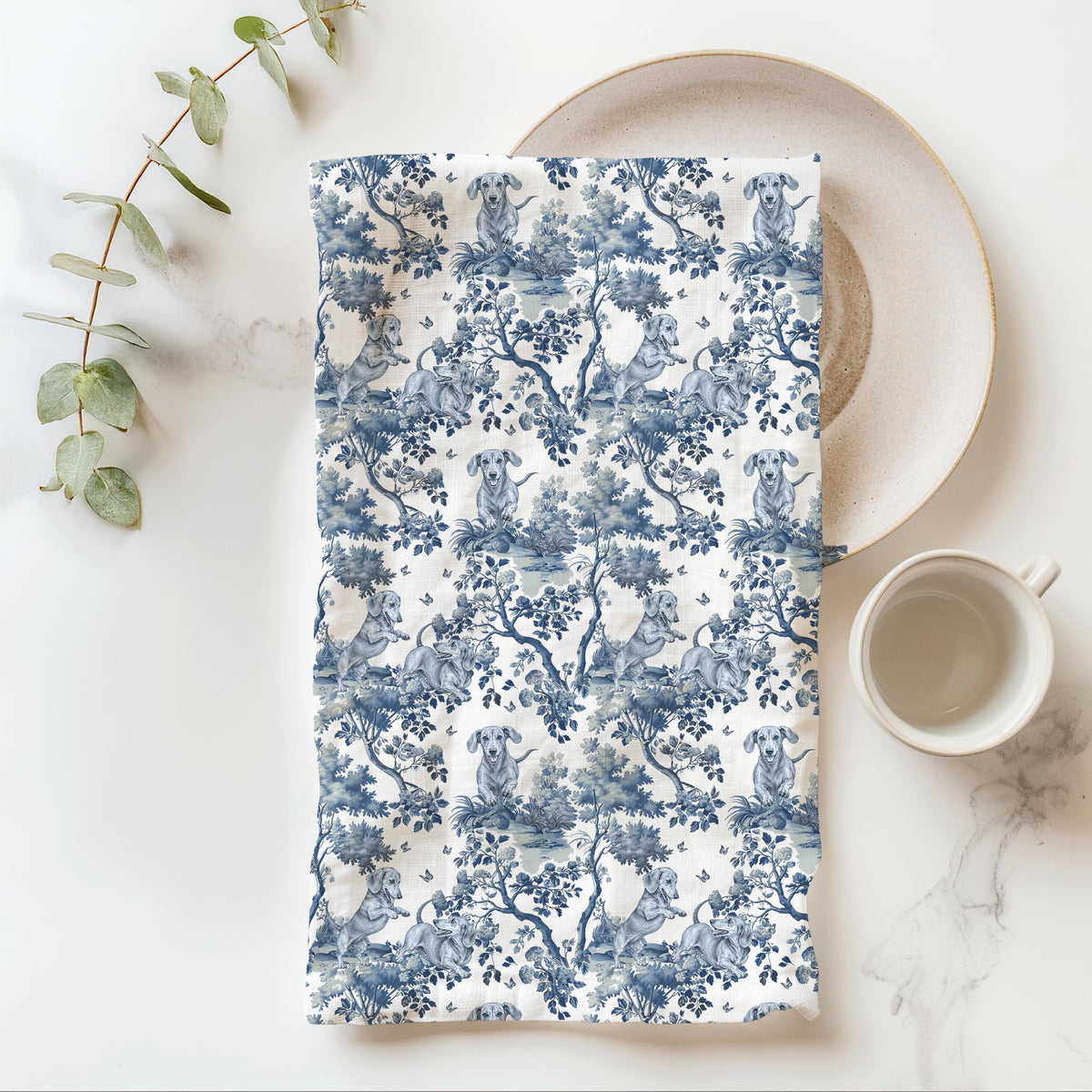 Dachshund Tea Towel with blue toile design, folded beside a plate and cup.