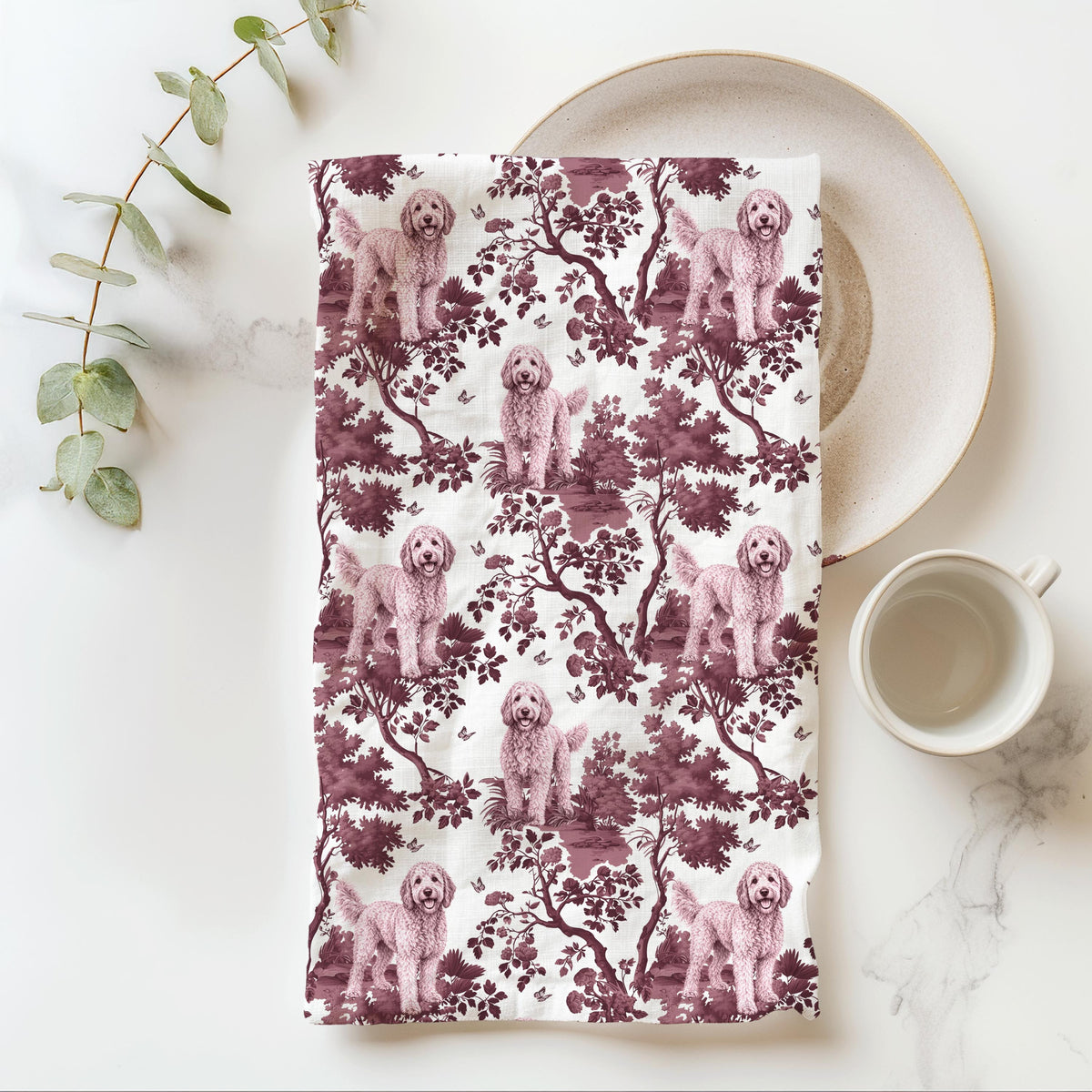 Goldendoodle Tea Towel with cranberry toile design, folded beside a plate and cup.