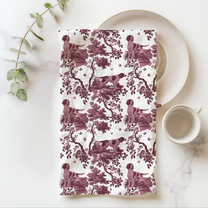 Irish Red and White Setter Tea Towel with cranberry toile design, folded beside a plate and cup.