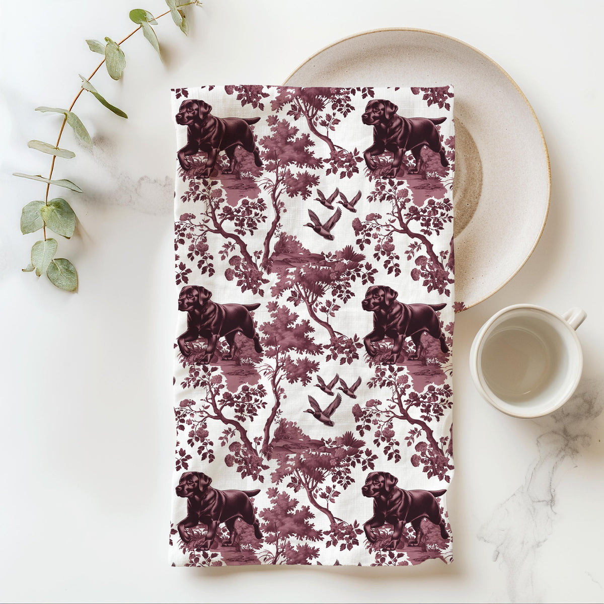 Black Labrador Duck Tea Towel with cranberry toile design, folded beside a plate and cup.