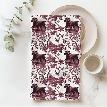 Black Labrador Duck Tea Towel with cranberry toile design, folded beside a plate and cup.