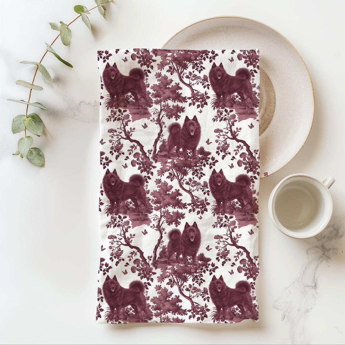 Schipperke Tea Towel with cranberry toile design, folded beside a plate and cup.