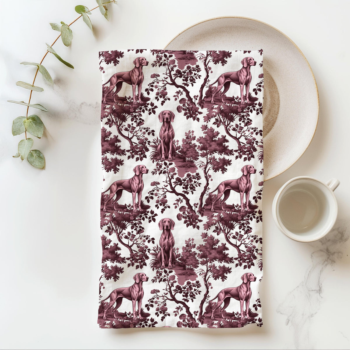 Weimaraner Tea Towel with cranberry toile design, folded beside a plate and cup.